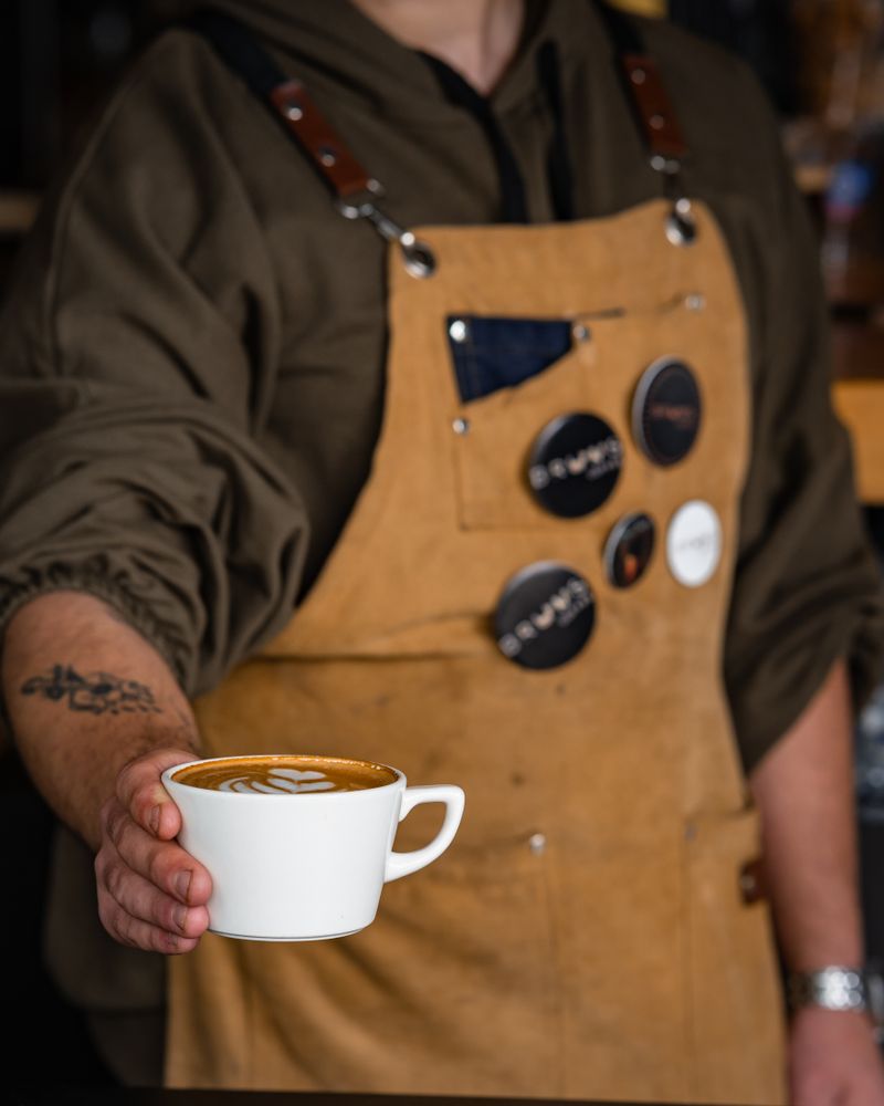 A barista in a tan apron holding out a freshly poured flat white